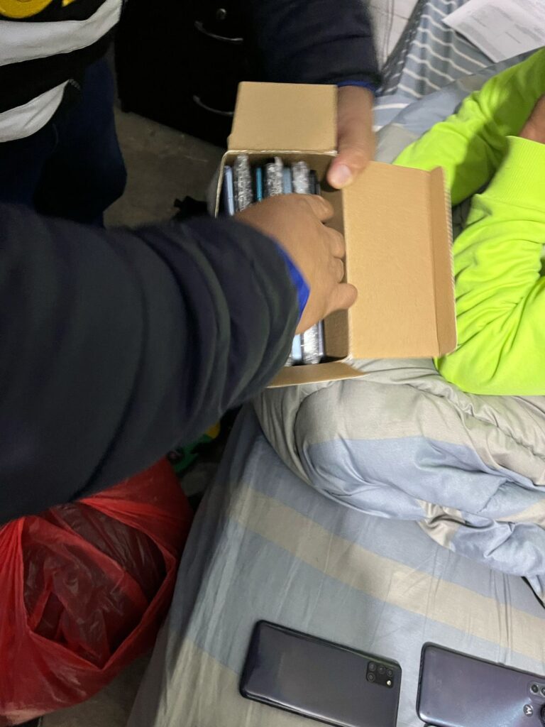 Una persona entrega una caja con varios artículos a otra persona en un entorno de hogar con teléfonos móviles y ropa visibles.